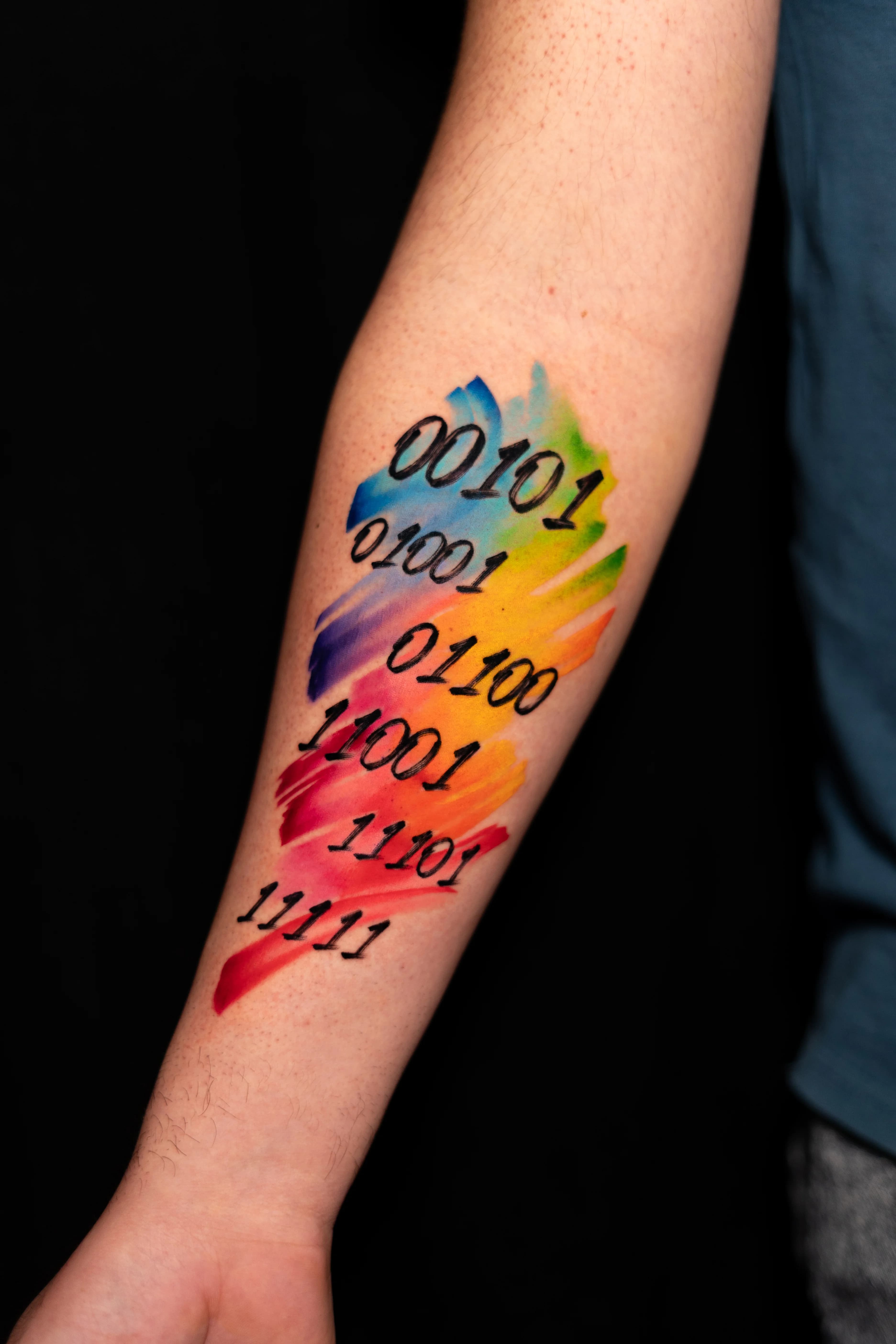 Un tatuaje en el antebrazo con números binarios superpuestos sobre un fondo colorido de estilo acuarela con tonos amarillos, azules, verdes, naranjas y rojos.
