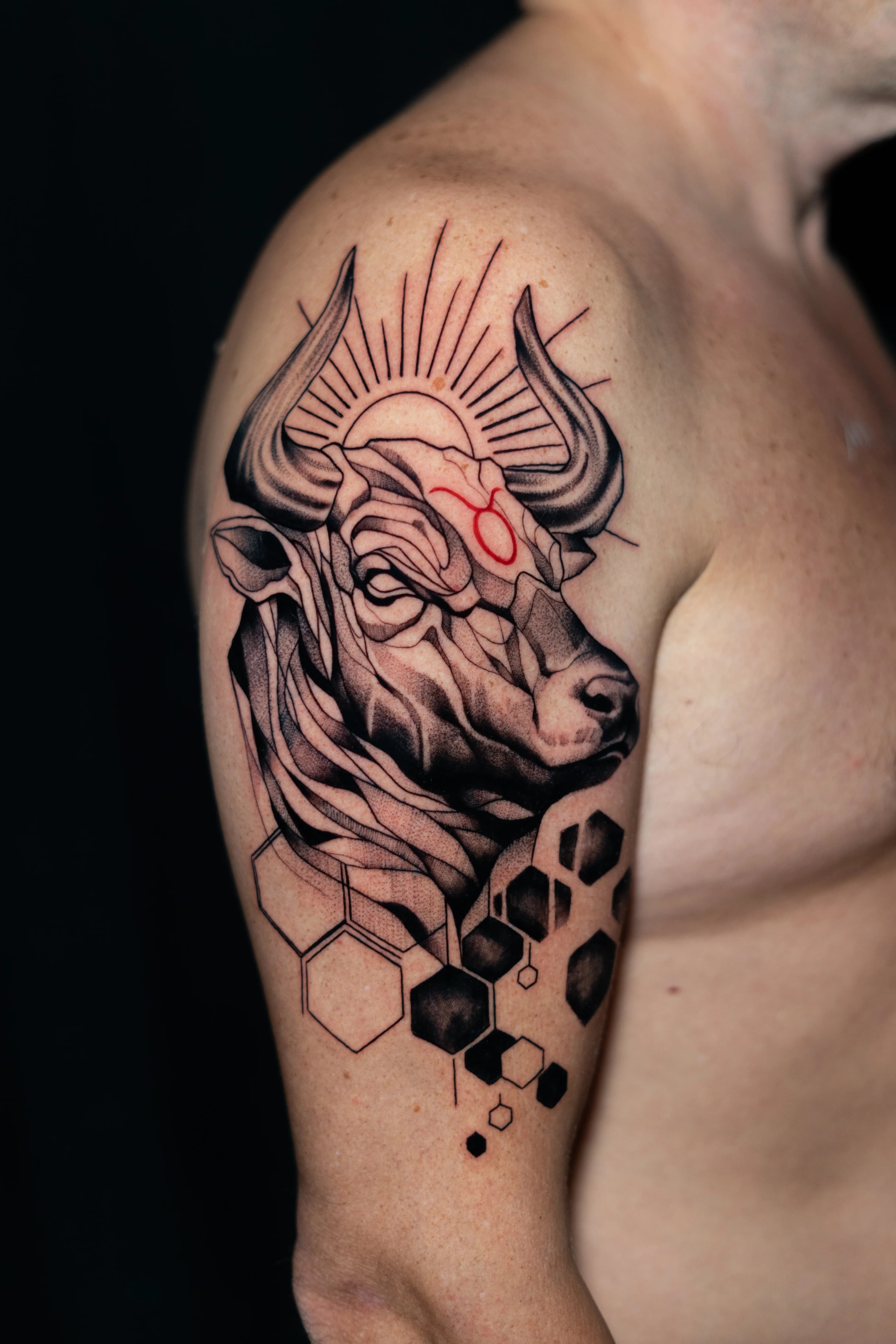 Un tatuaje detallado de una cabeza de toro adornada con rayos y hexágonos geométricos, con un símbolo del zodíaco rojo en el brazo de una persona.