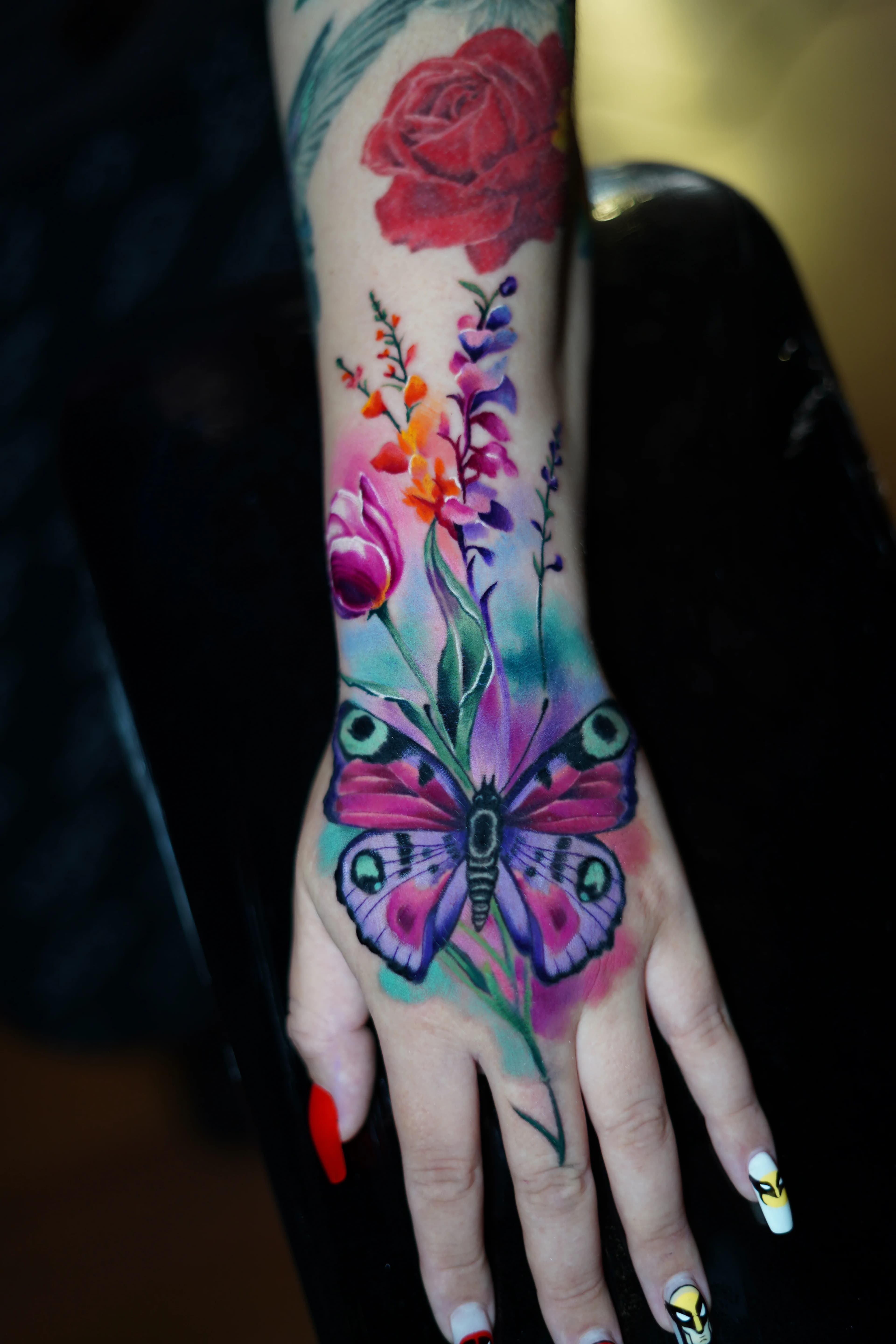 Un tatuaje vibrante de una gran mariposa morada rodeada de flores de colores en una mano con uñas rojas.