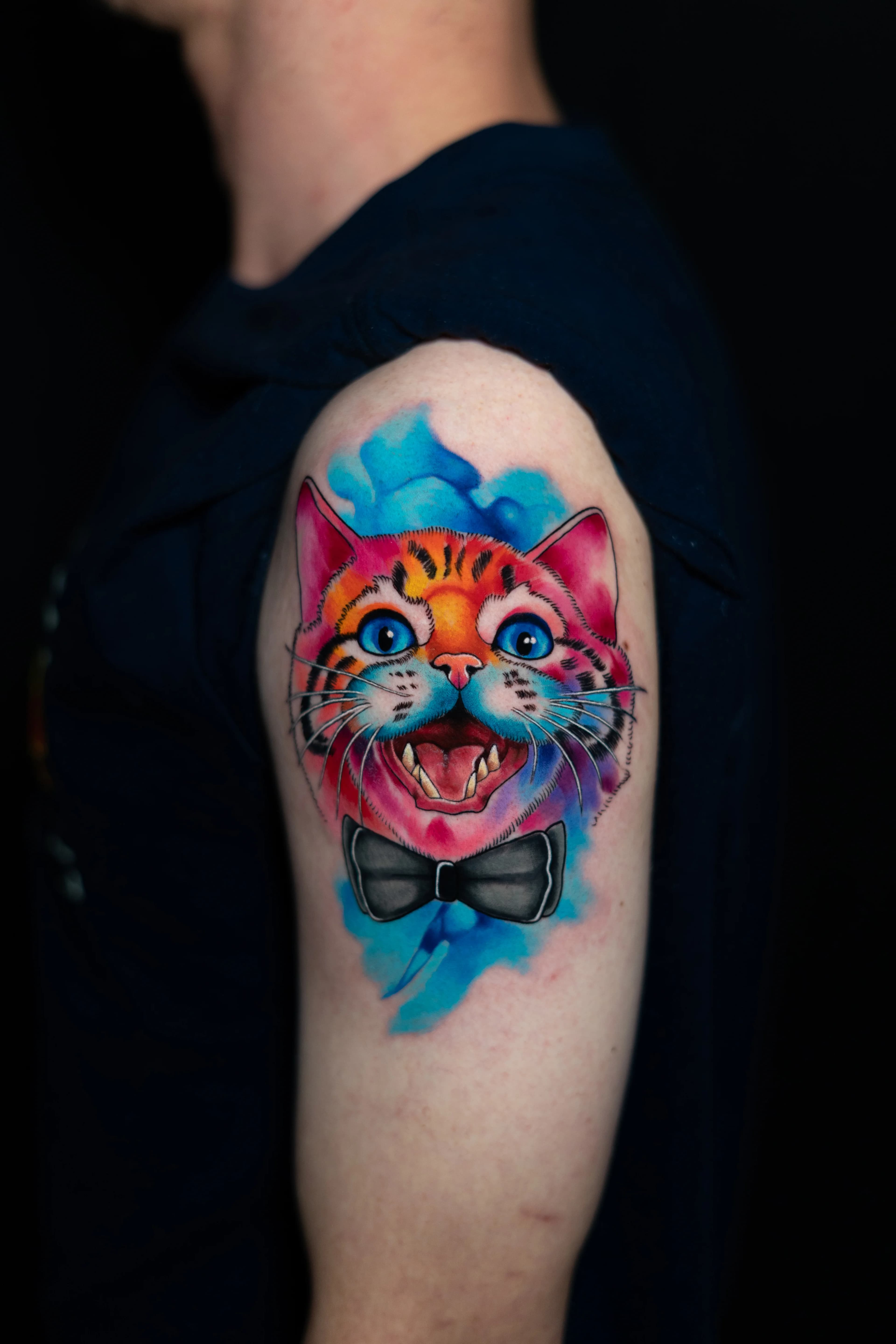 Un tatuaje vibrante de un gato sonriente con una pajarita, con salpicaduras de acuarela de colores en el fondo, en la parte superior del brazo de una persona.