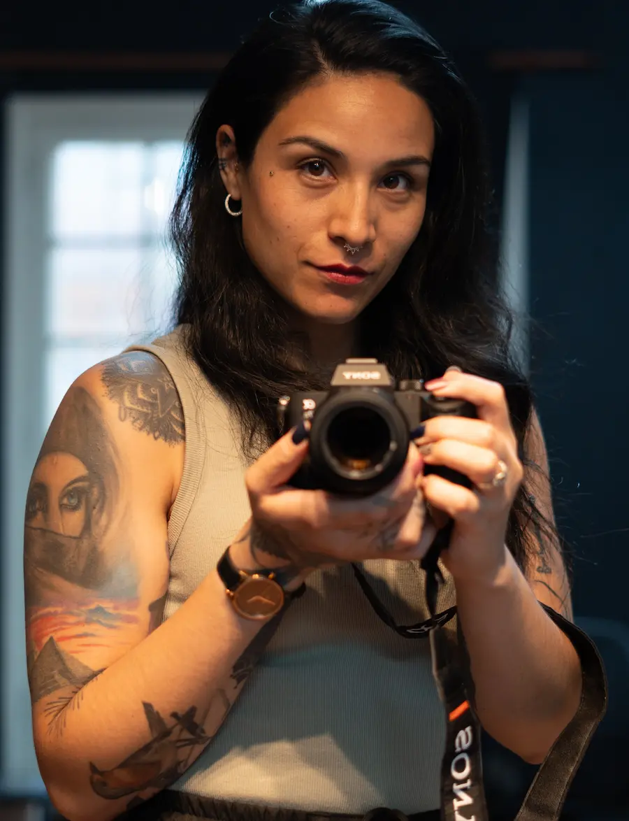 Aramoonlight sostiene una cámara, exhibiendo tatuajes detallados en el brazo y vistiendo un atuendo elegante, en una habitación suavemente iluminada.